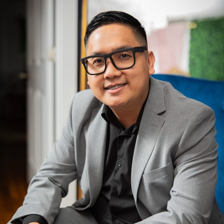 Jay Dural Smiling man in glasses wearing a gray suit and black shirt, sitting on a blue chair.